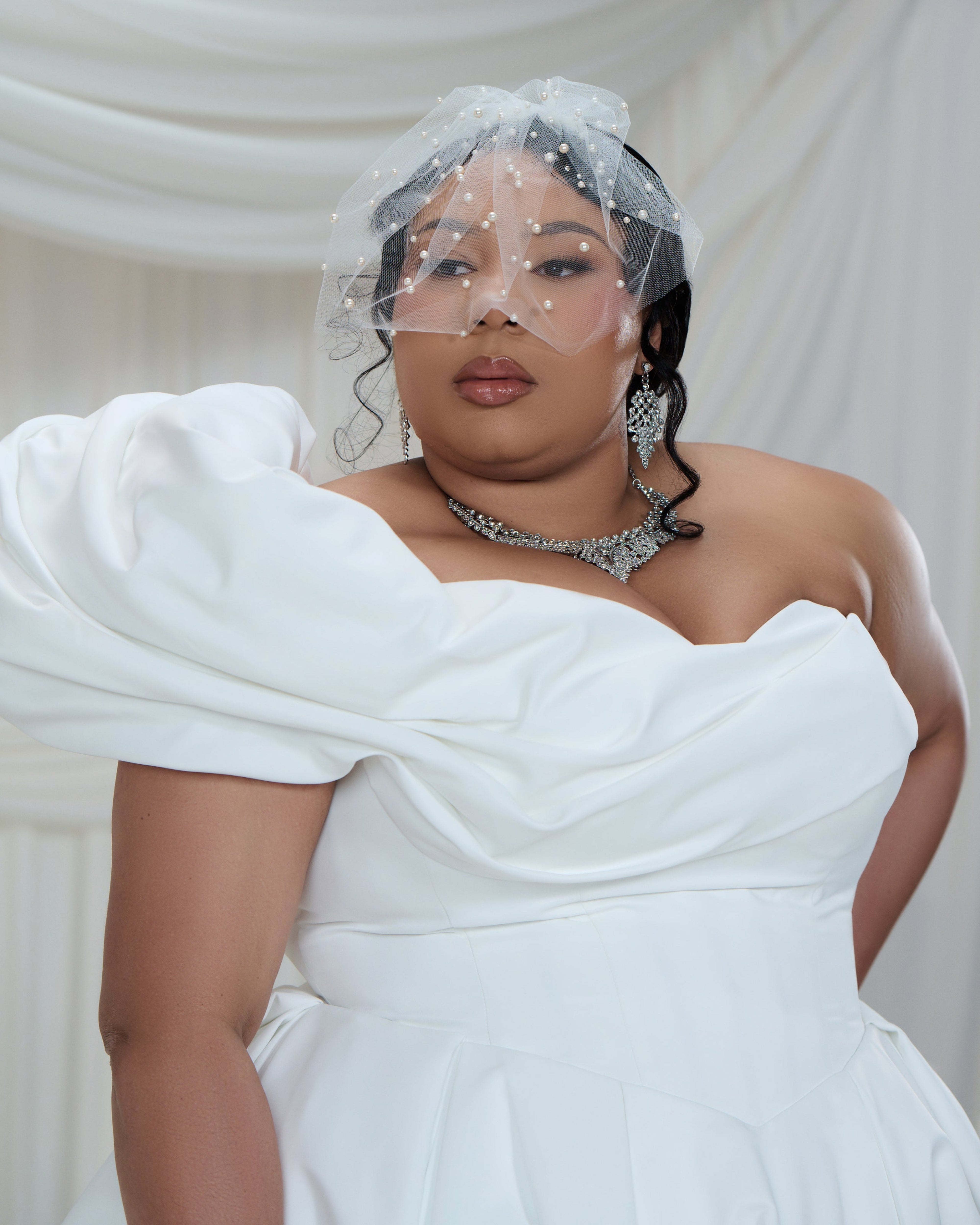 A plus-size model wearing a white off-the-shoulder wedding dress with dramatic puffed sleeves, a sheer embellished veil, and silver jewelry, posed against a draped white fabric backdrop.
