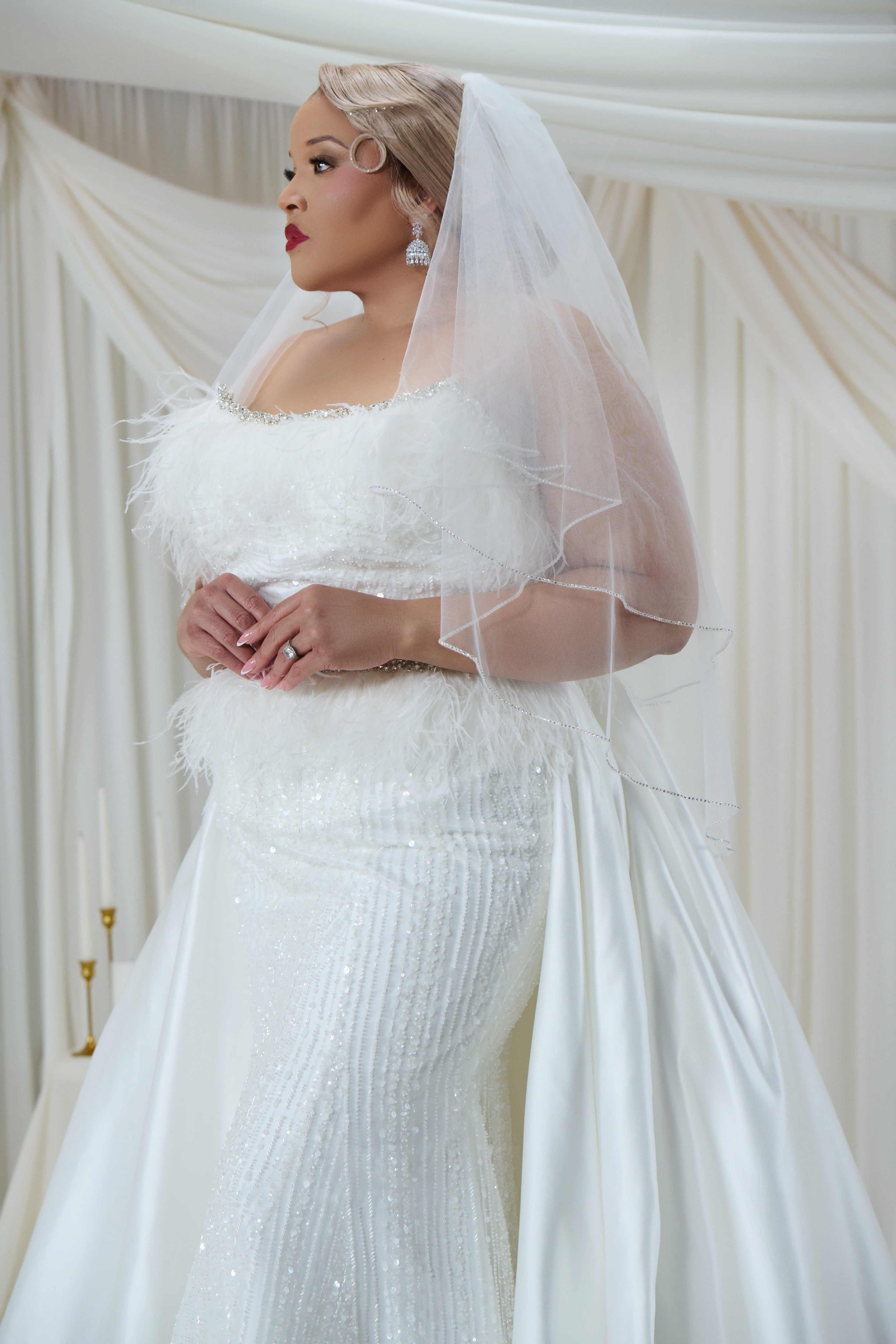 A woman wearing a white off-the-shoulder wedding dress with feathered detailing, a sheer long veil, and a fitted sequined mermaid-style skirt, posed against a draped cream fabric backdrop.