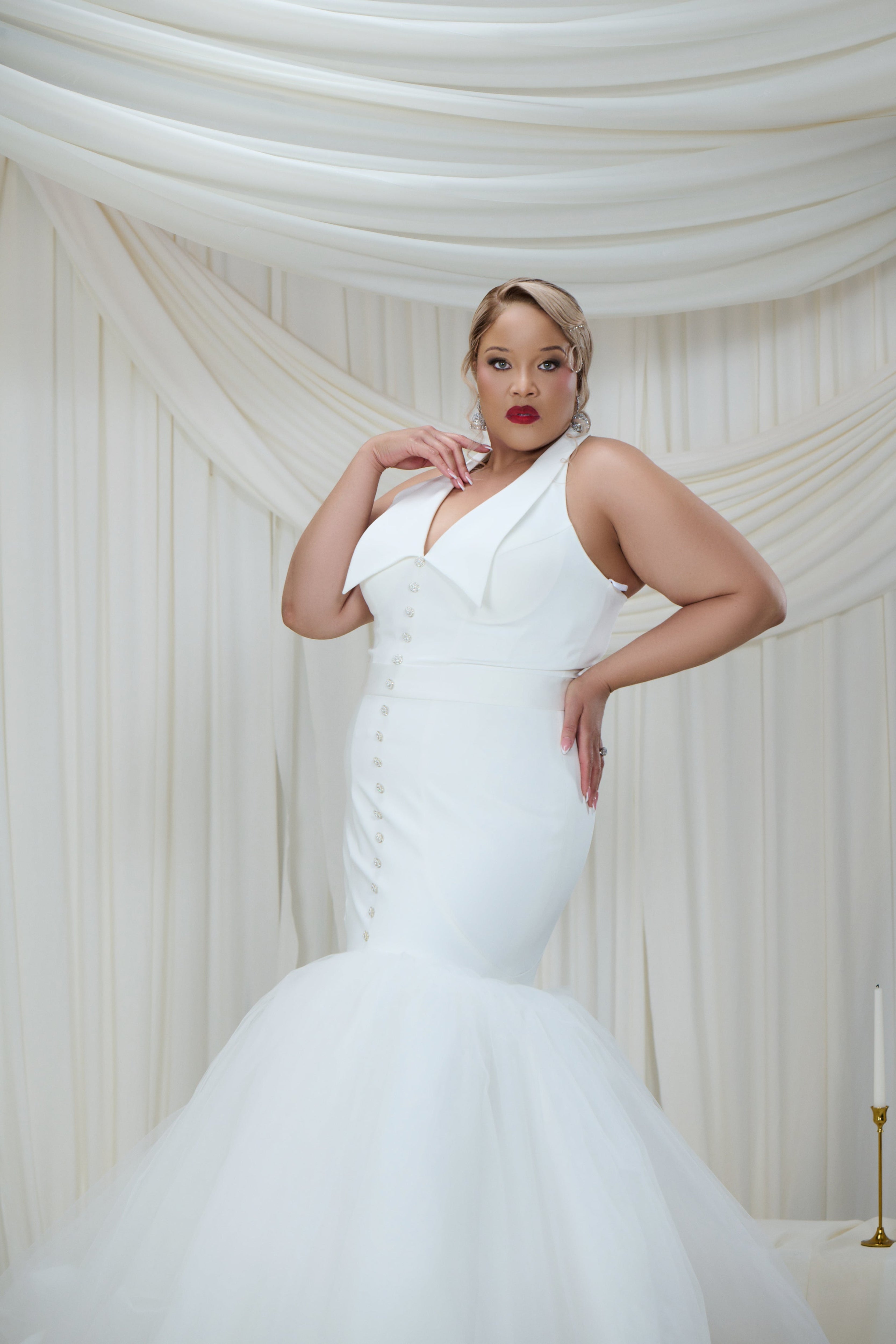 A woman modeling a white sleeveless mermaid-style wedding gown with a structured corset bodice and full tulle skirt, posed against draped cream fabric curtains.