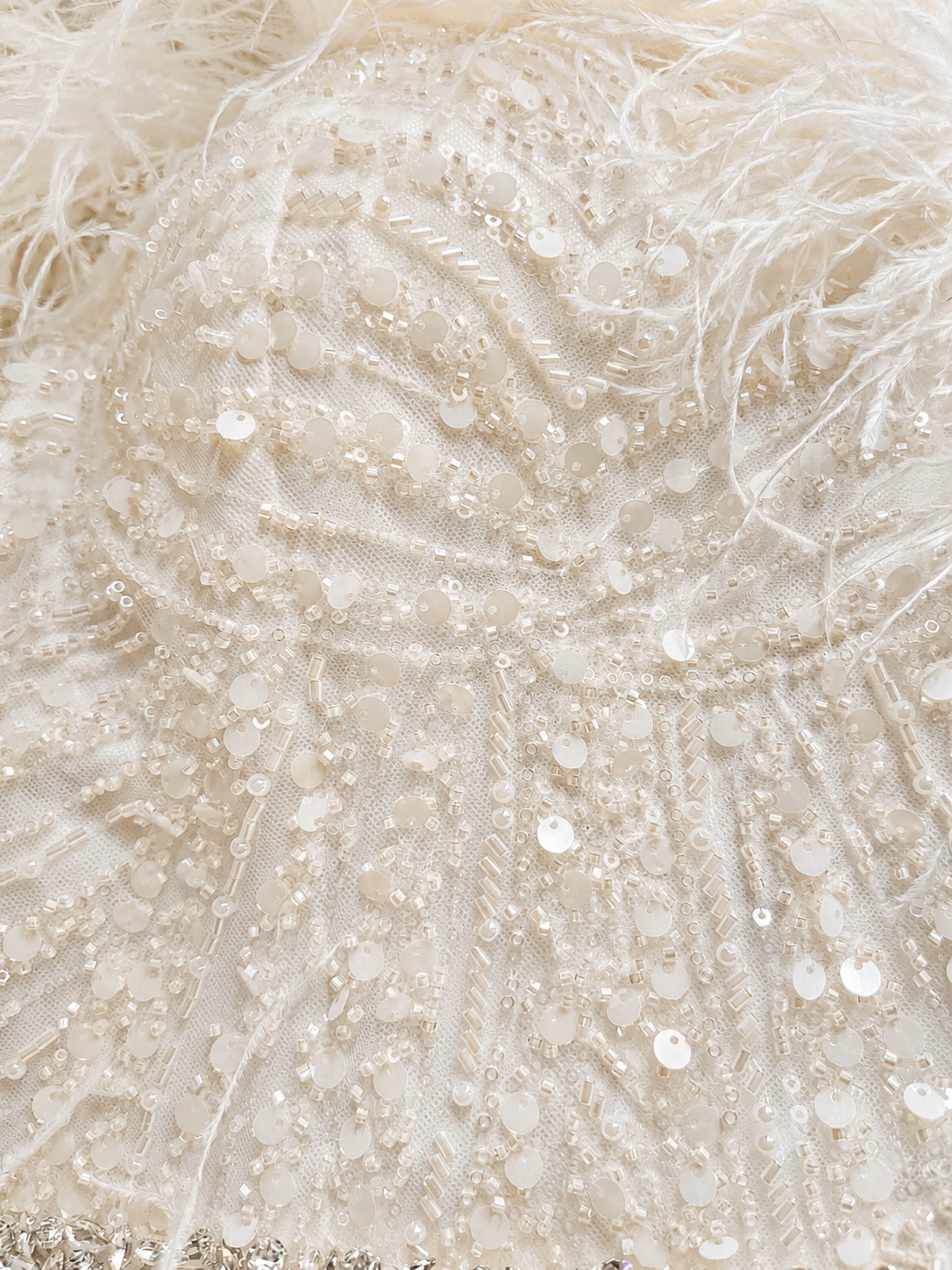 Close-up of a cream-colored embellished fabric with feathers, sequins, beads, and mesh detailing.