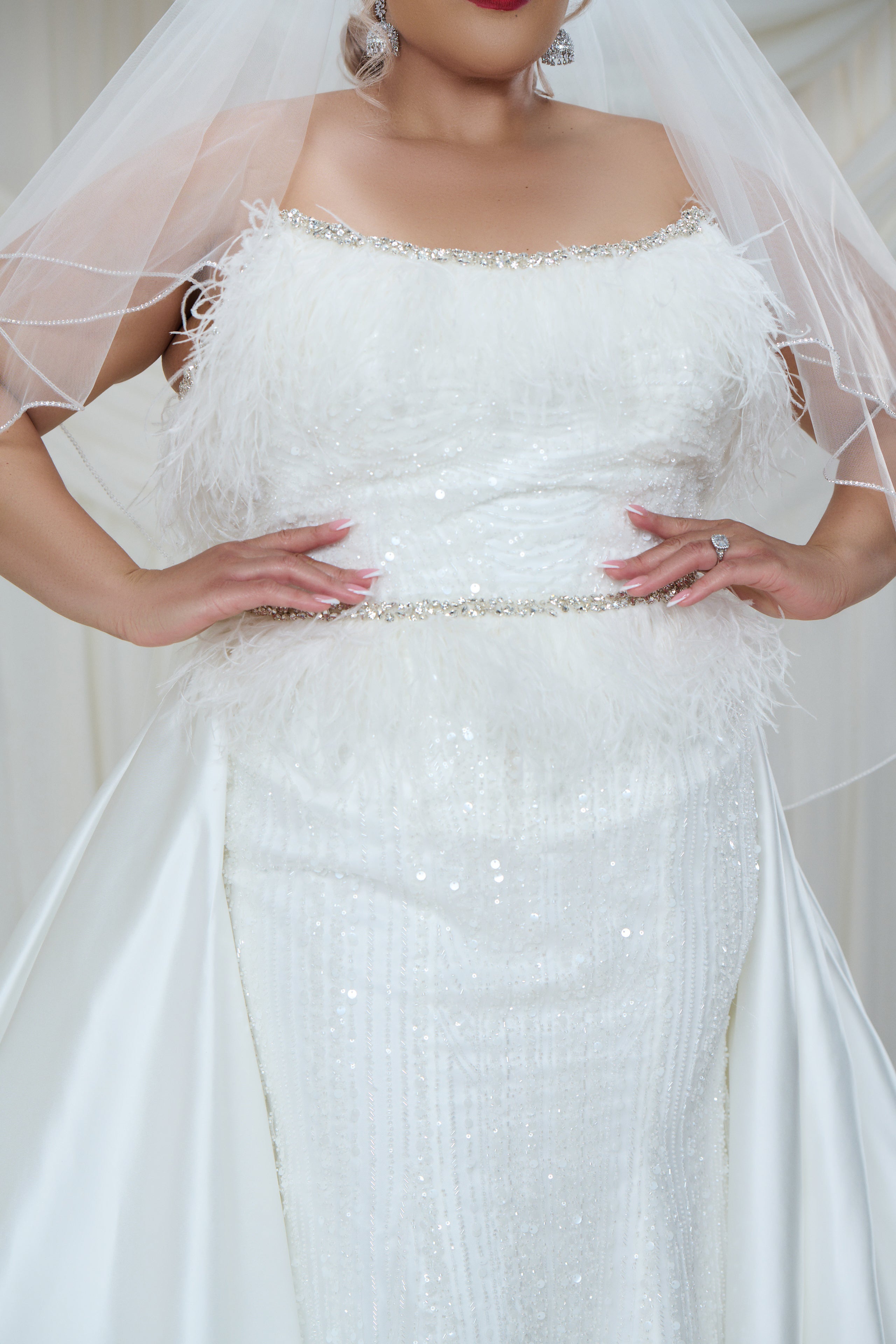 A close-up of a white off-the-shoulder wedding dress with feathered trim, a beaded waistband, and a sheer veil, shown on a model.