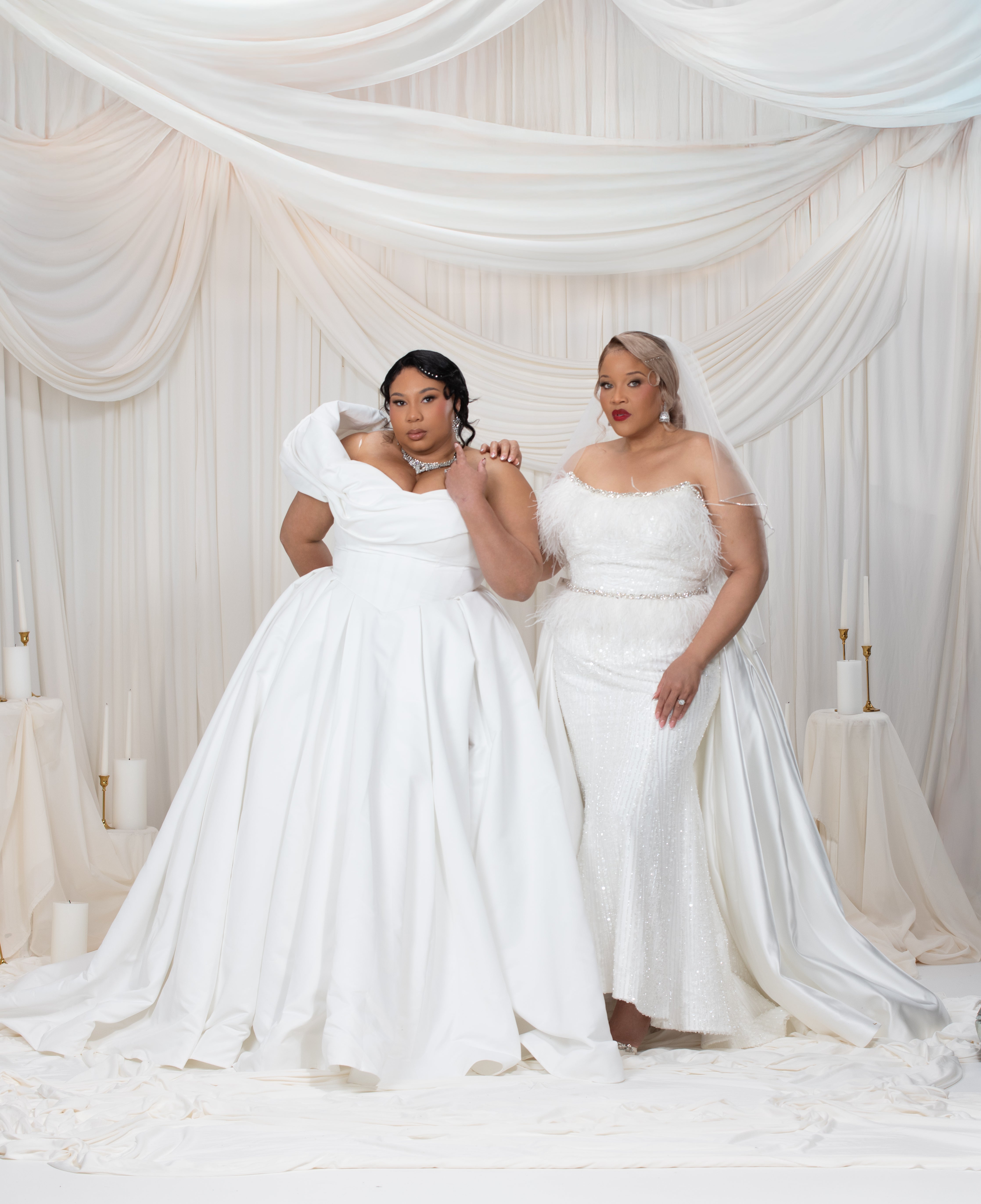 Two women in white wedding dresses standing in front of a draped white curtain.